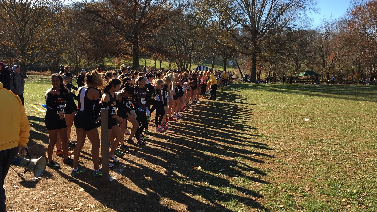 A line of high school girls in team uniforms stands at the start line of a cross-country race on a grassy field while officials check the line.