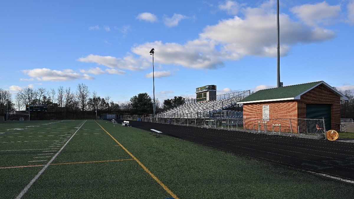 An empty high school stadium on a sunny morning: artificial turf field, a running track with hurdles stacked beside it, aluminum bleachers and a brick equipment shed.