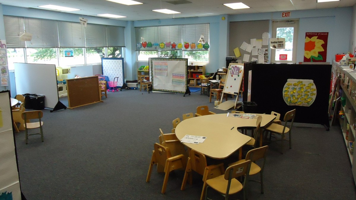 An empty preschool classroom with small tables and chairs, toys on shelves and children's art on the walls.