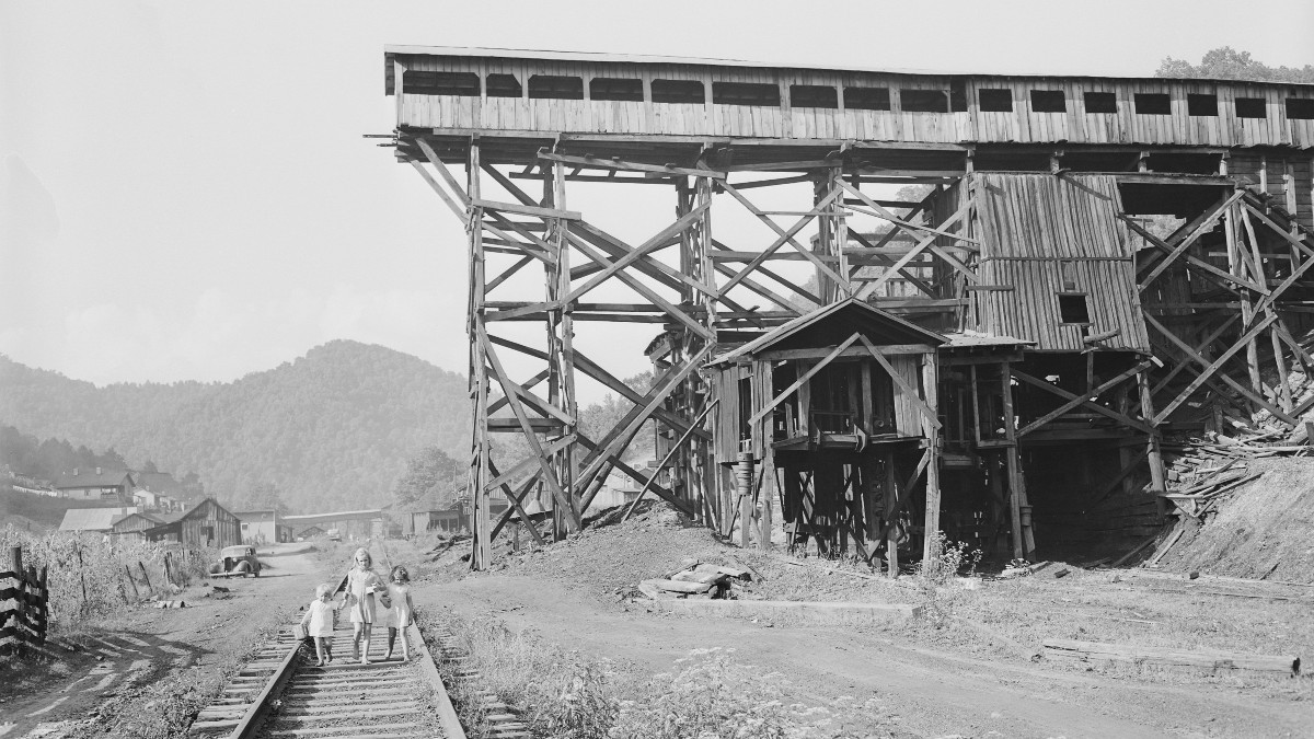 Black-and-white photo of a large, weathered wooden coal tipple on timber stilts beside a railroad track in a Kentucky hollow, with three children walking on the track and wooded hills and miners' houses in the background.