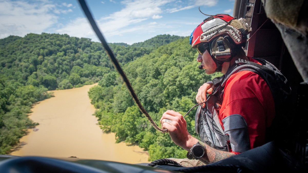 A Kentucky Air National Guard airman in a red helmet looks out of a helicopter over a muddy, flooded river winding between forested hills in eastern Kentucky.