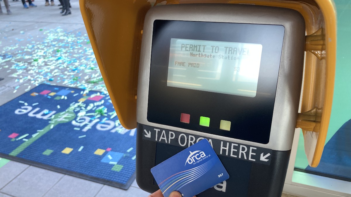 A rider holds a blue ORCA card to a yellow-hooded card reader at Northgate light rail station; the screen reads Permit to Travel, Fare Paid.