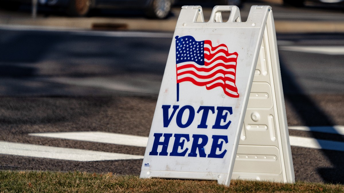 A white sandwich-board sign with an American flag and the words 'Vote Here' outside a polling place, with parked cars behind.