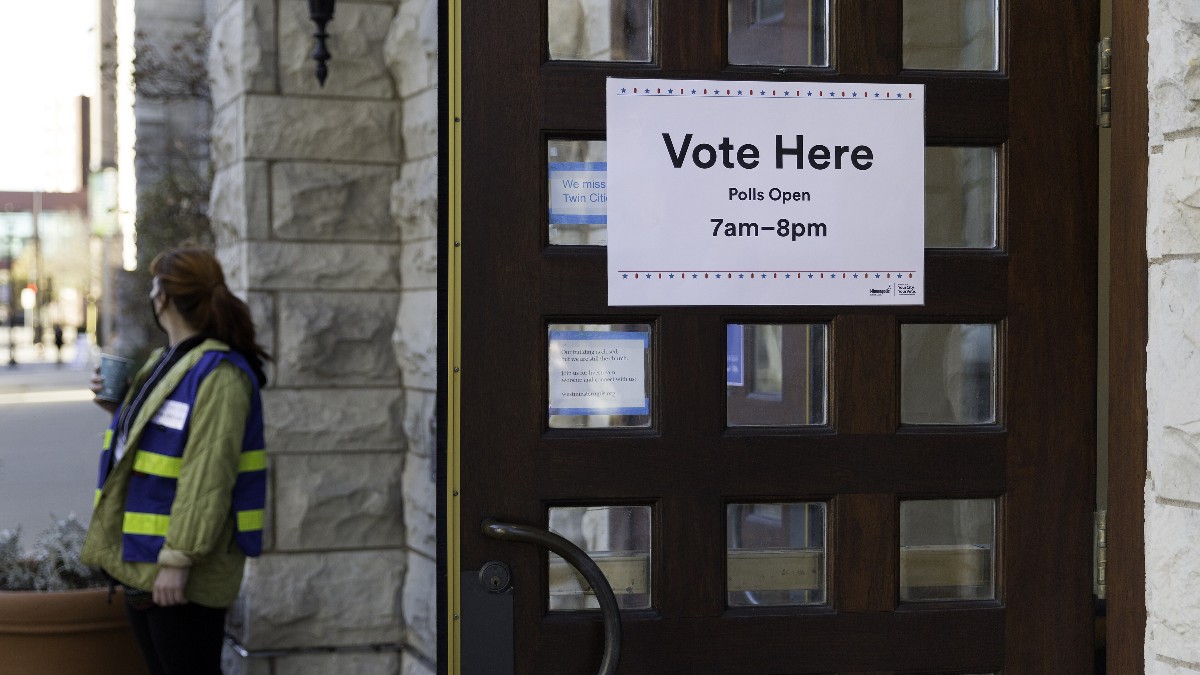 A 'Vote Here, Polls Open 7am-8pm' sign on a wooden door at a polling place, with an election official in a safety vest walking past.
