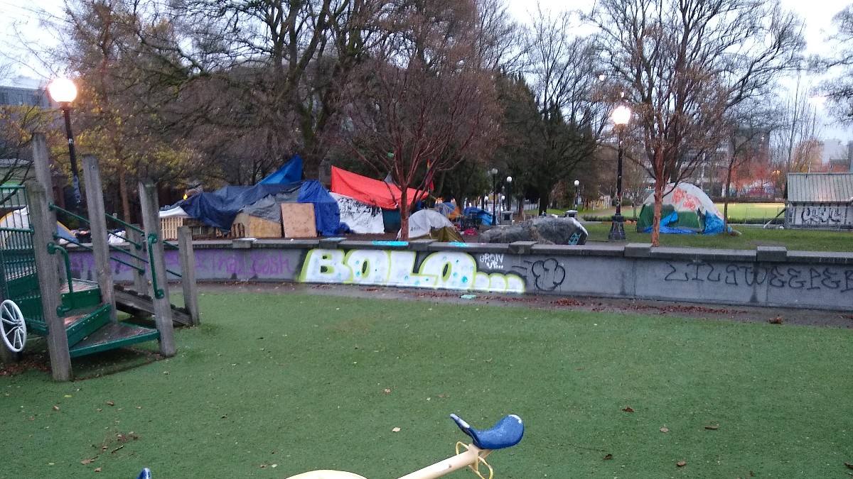 Tents and tarps lined up along a low graffiti-covered wall in a Seattle park, behind an empty children's playground with a seesaw.