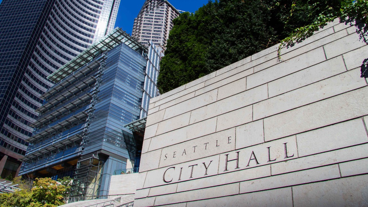 Low-angle view of Seattle City Hall's glass facade beside a stone wall carved with the words Seattle City Hall, with downtown towers and a blue sky behind.