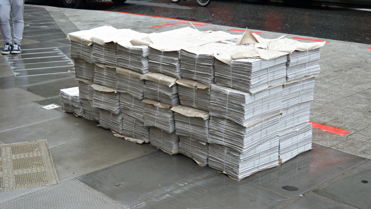 Bundles of newspapers stacked on a city sidewalk, with a bus and a cyclist on the street behind them.