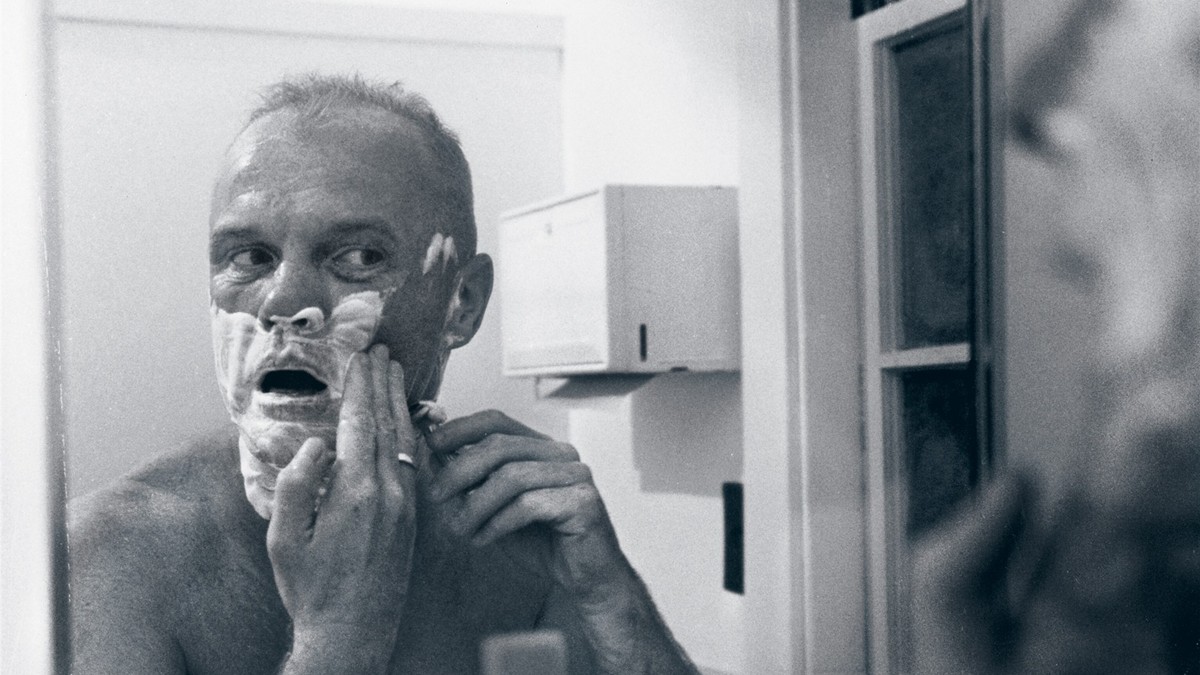 Black-and-white photograph of astronaut John Glenn shaving his face with a safety razor at Cape Canaveral in 1962.