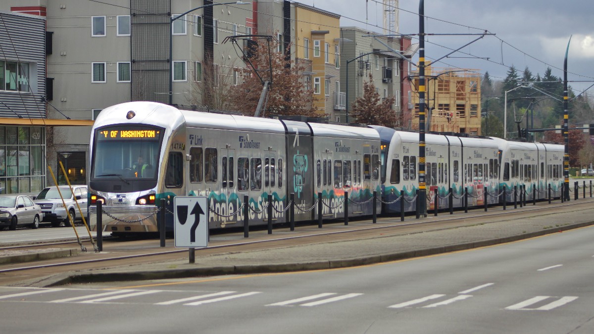 A three-car Sound Transit Link light rail train running beside a street lane on Martin Luther King Jr. Way South in Seattle, with parked cars at left.