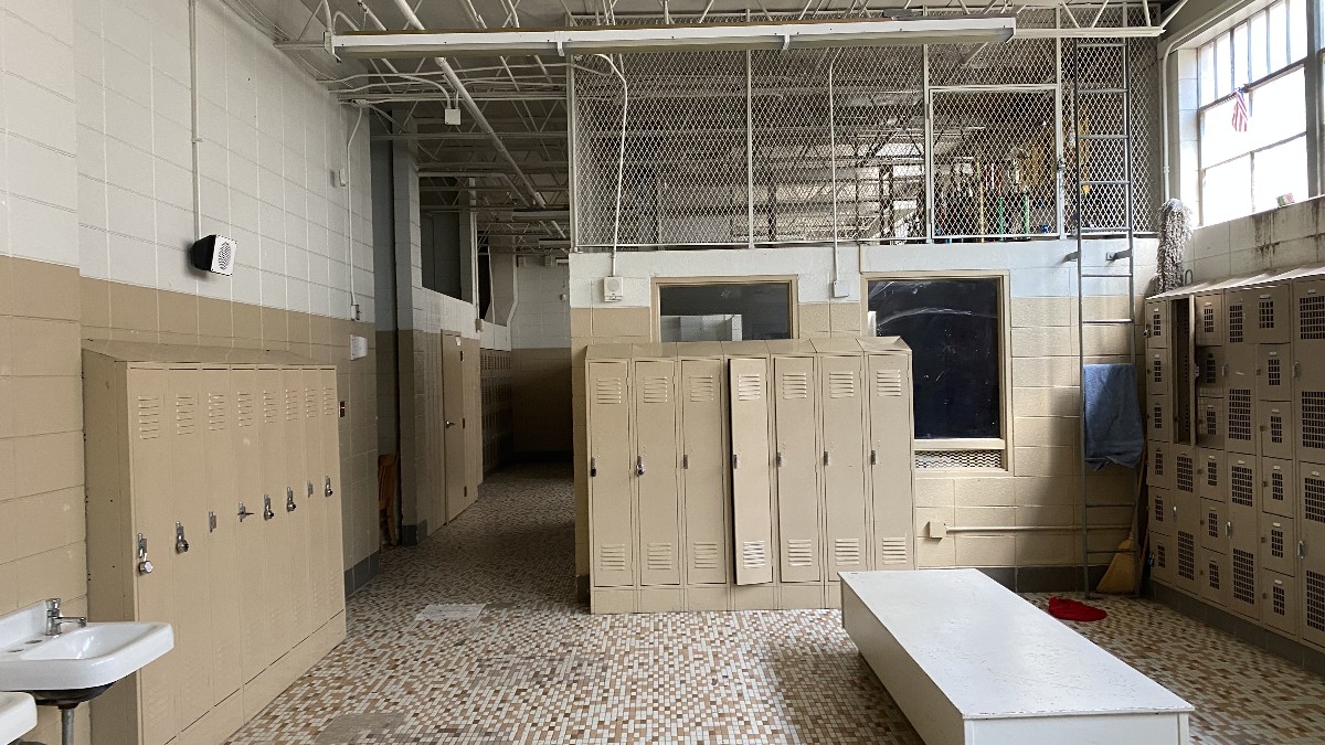 An empty high school locker room with tan metal lockers, white benches and a tiled floor.