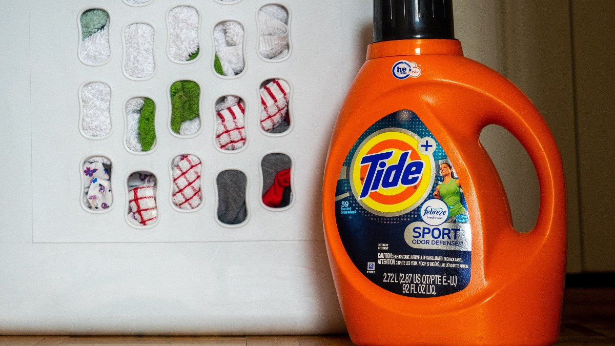An orange bottle of Tide liquid laundry detergent standing on a wooden floor beside a white wall panel.