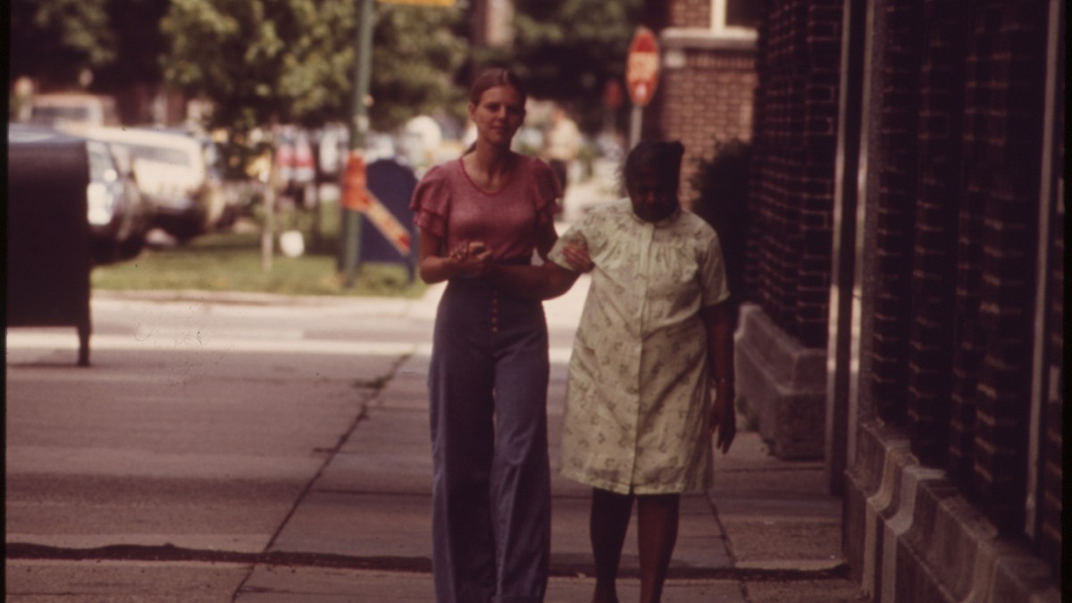 A young woman steadies an older woman by the arm as they walk along a city sidewalk.