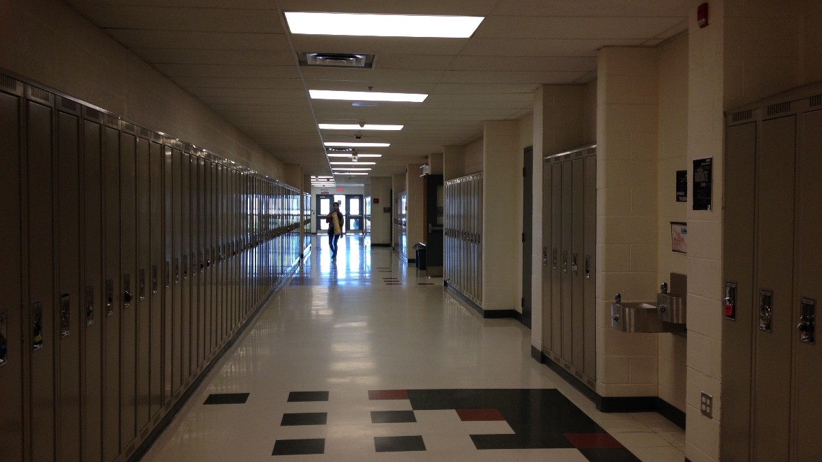 A long, mostly empty school hallway lined with gray lockers, with a single distant figure walking toward the doors at the far end.