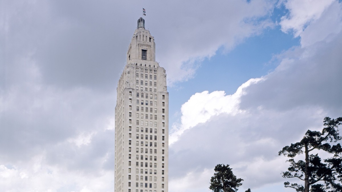 The Louisiana State Capitol tower in Baton Rouge rising above trees and a lawn under a cloudy blue sky.