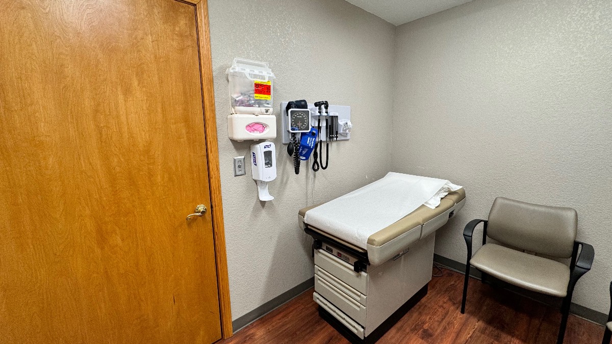 An empty medical exam room with a paper-covered exam table, a wall-mounted blood pressure gauge, a chair and a closed wooden door.
