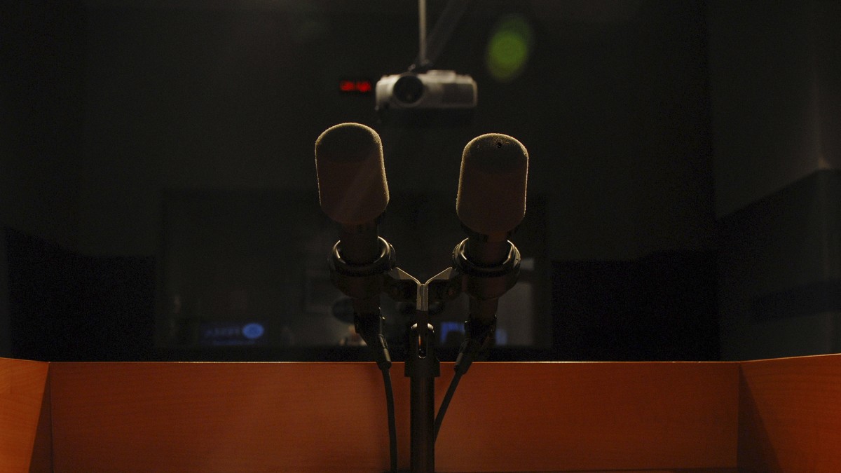 Two foam-covered microphones on a stand at an empty wooden podium in a dim press briefing room.