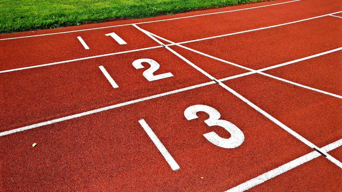 Red running track with white lane lines and large lane numbers 1, 2 and 3 painted at the start line, grass at the top edge.