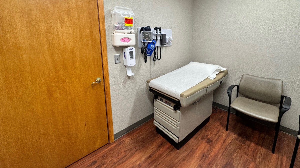 An empty examination room in a doctor's office, with a paper-covered exam table, a chair, and wall-mounted blood pressure and otoscope equipment.