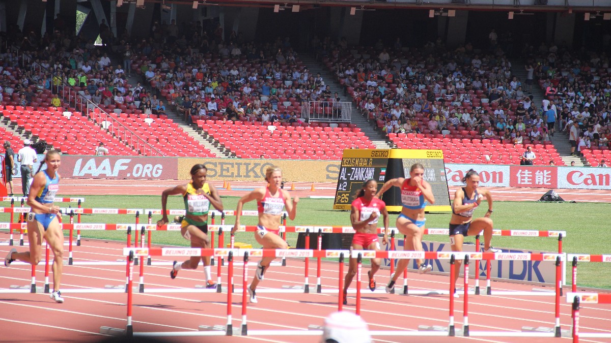 Six women clear hurdles in a 100 metres hurdles heat on a red track in front of a stadium crowd.
