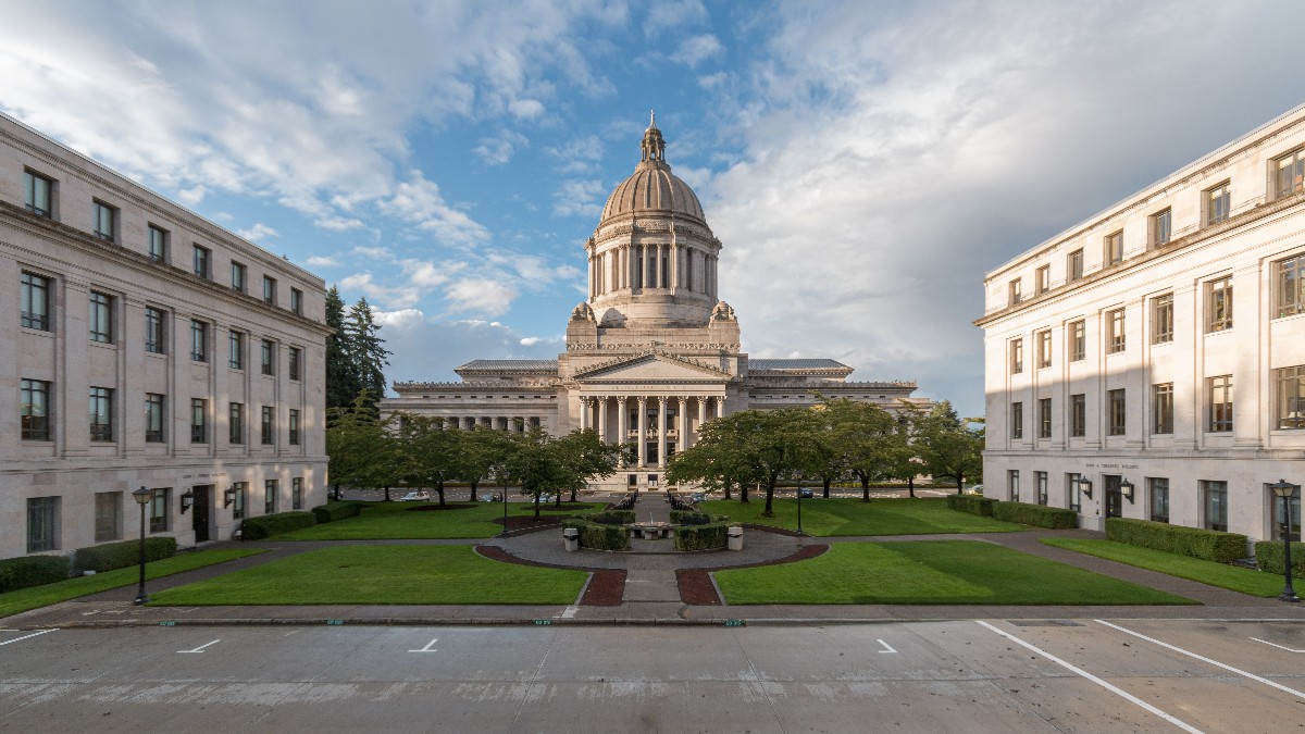 The domed Legislative Building of the Washington State Capitol in Olympia, seen across a lawn between two stone office buildings under a partly cloudy sky.